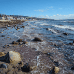 Seaglass hunting on rocky beach with waves and coastal village in background.